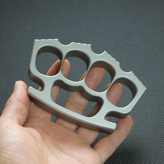 Hand holding silver four-hole knuckle duster against black textured mat, palm-side view.