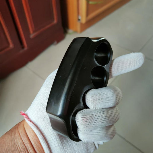 A black knuckle duster shown at a three-quarter angle in a white-gloved hand, displaying its thick side edge and contoured front face, on a blurred indoor background.