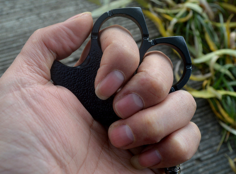 Black two-finger knuckle worn on a closed fist outdoors with a wooden deck and plants behind