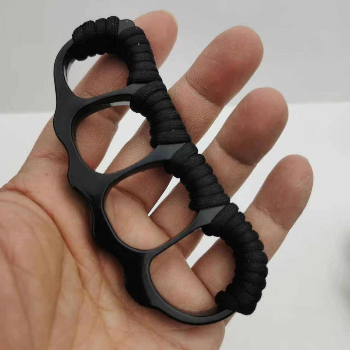 Matte black knuckle duster resting in an open palm, black paracord coils along the upper edge