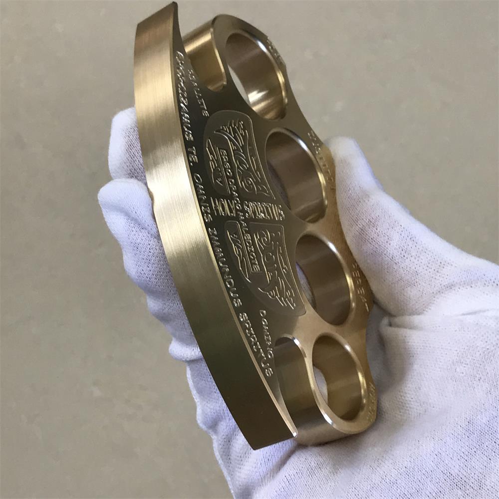 Brass knuckle duster held in a gloved hand showing the curved bottom edge lettering and satin side walls