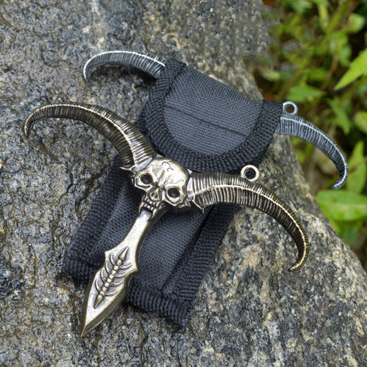 Bronze sheep skull pendant with curved ribbed horns resting on a black nylon sheath with a silver gray horn piece behind on rock