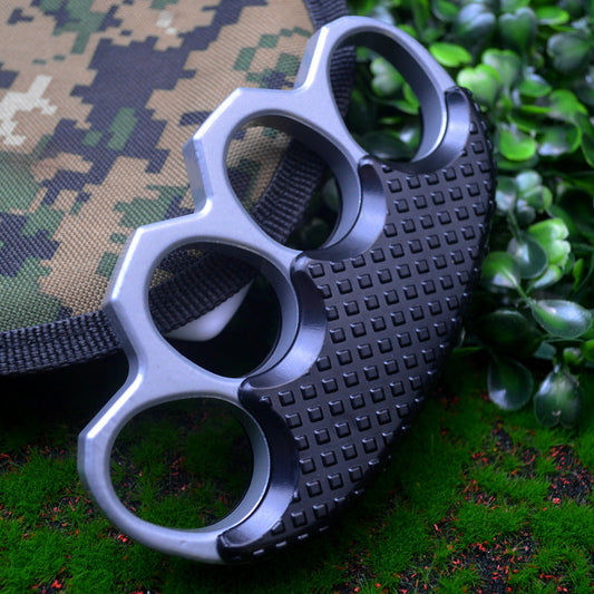 Silver gray knuckle duster with a black studded grip panel leaning against a camouflage pouch and black webbing strap on grass