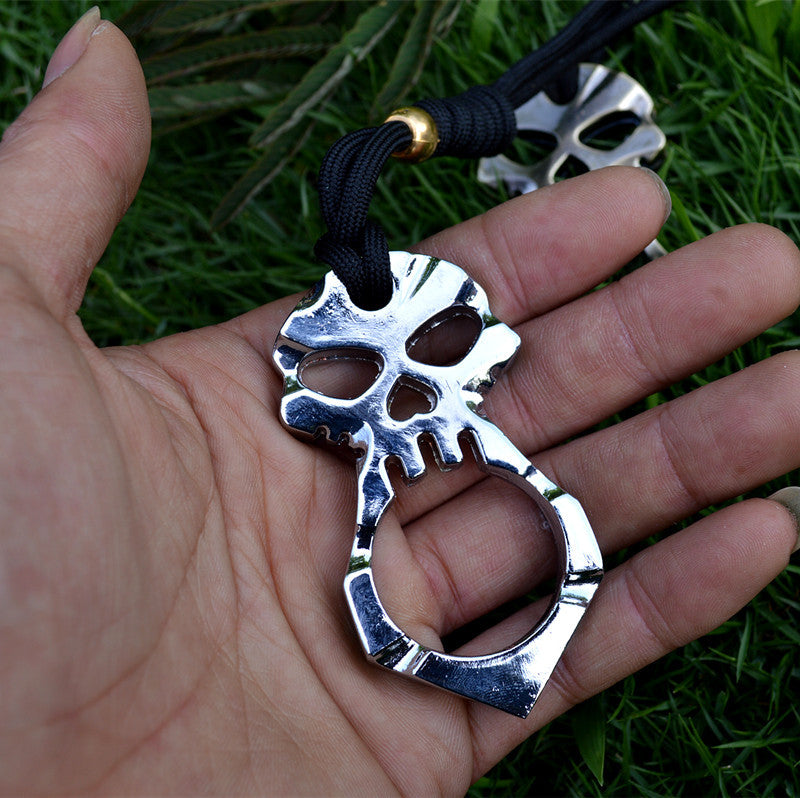 A hand holds the silver skull knuckle duster against green grass outdoors, its heart-shaped hole and eye sockets in view above the single finger ring, with a second lanyarded piece blurred behind.
