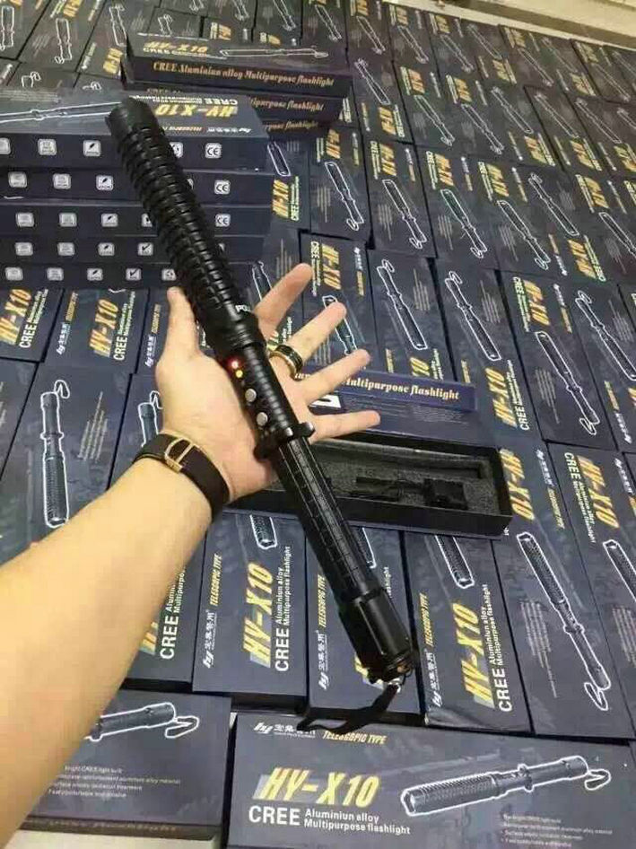 Hand holding the black baton-style stun gun with ribbed expandable head, knurled grip and wrist strap, retail boxes stacked in the background