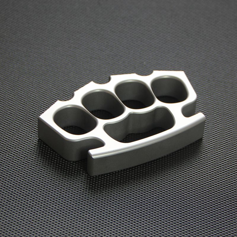 Silver knuckle duster at slight angle on black textured mat, showing all four finger holes.