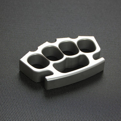 Silver knuckle duster at slight angle on black textured mat, showing all four finger holes.