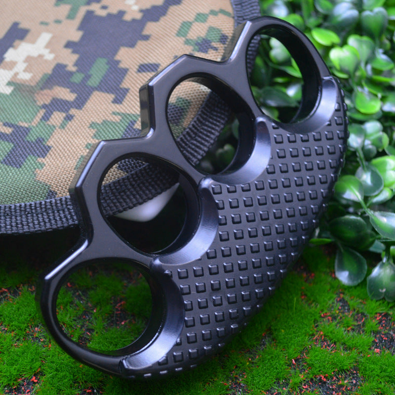 All black knuckle duster in close-up against a camouflage pouch on grass, four oval finger holes and studded grip panel