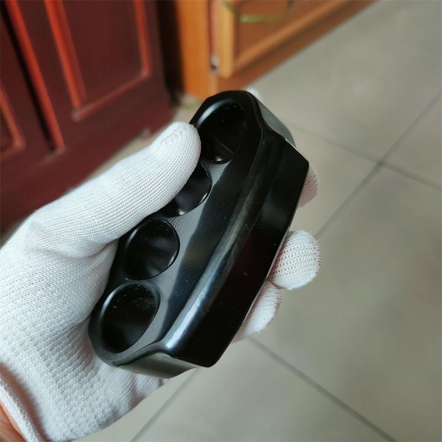 A glossy black knuckle duster held at an angle in a white-gloved hand, thumb on top, with rounded edges and four recessed holes, on a blurred tiled background.