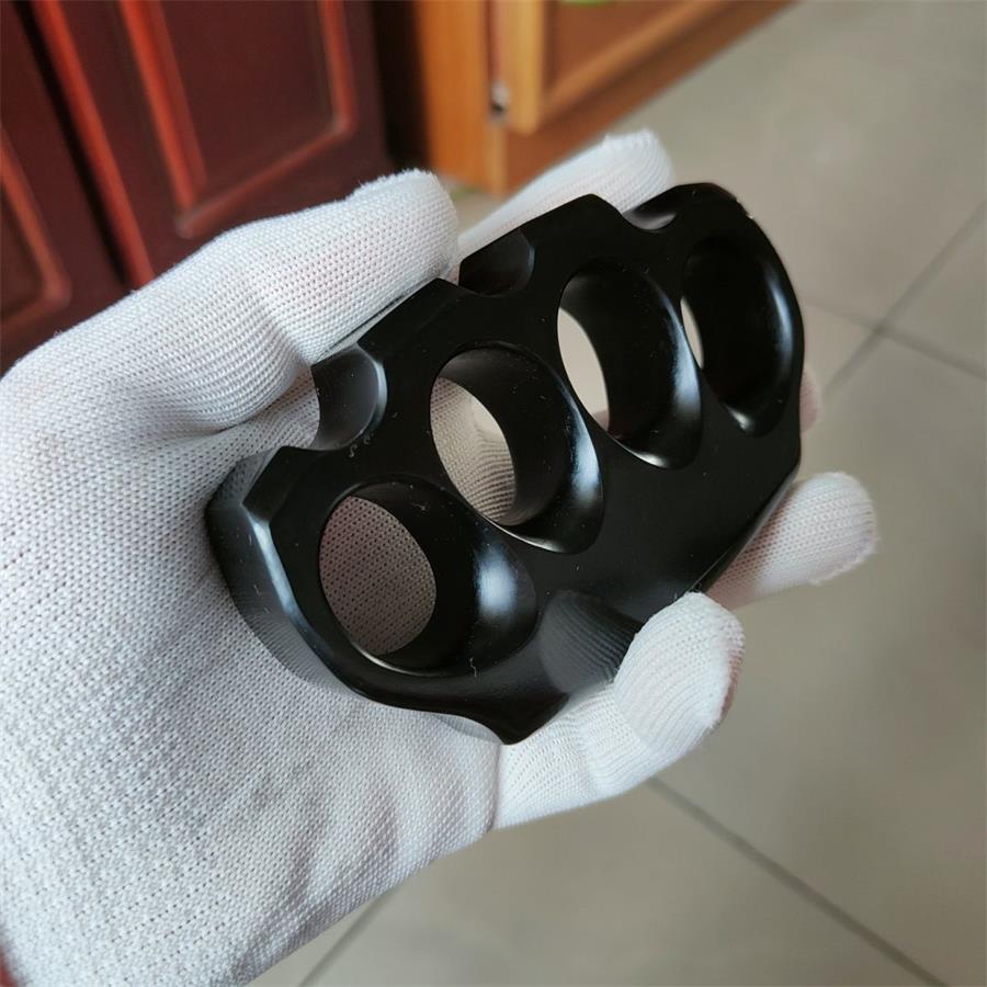 A close-up of a black knuckle duster held in a white-gloved hand, showing the rounded finger-hole edges and contoured top grooves, on a blurred wooden background.