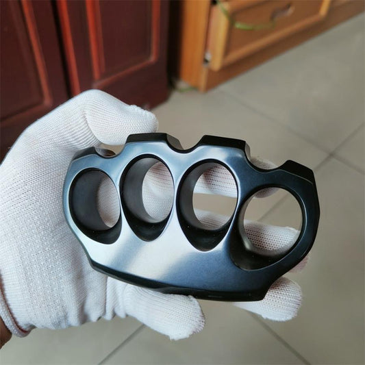 A glossy black knuckle duster with a blue-gray sheen, shown face-on in a white-gloved hand, four finger holes in a row with scalloped knuckle bumps, on an indoor tiled background.