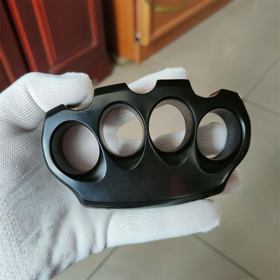 A matte black knuckle duster held flat in a white-gloved hand, four graduated finger holes and a scalloped top edge, on a blurred indoor background.