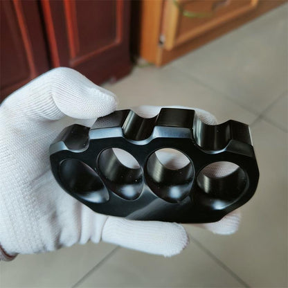 A glossy black knuckle duster held palm-up in a white-gloved hand on a beige tiled floor, with blurred wooden furniture in the background, indoors.