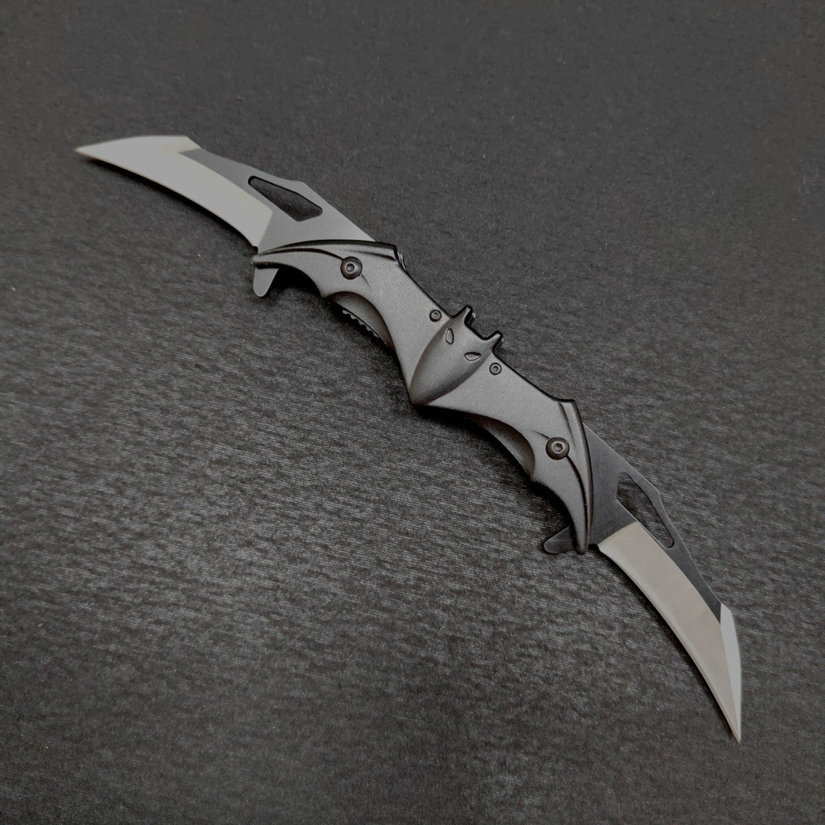A fully open black bat-shaped double folding knife on a dark gray surface, with two drop-point blades extended one at each end and a central bat body showing ears, eyes, spread wings and slotted edges.