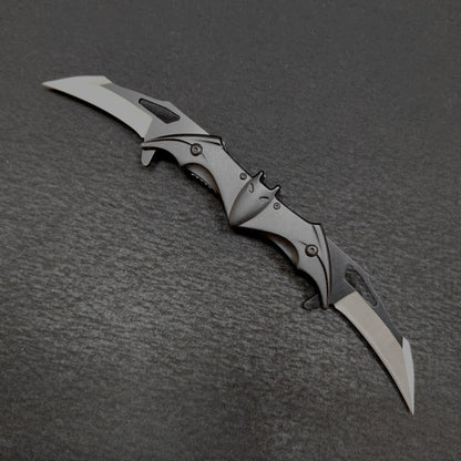 A fully open black bat-shaped double folding knife on a dark gray surface, with two drop-point blades extended one at each end and a central bat body showing ears, eyes, spread wings and slotted edges.