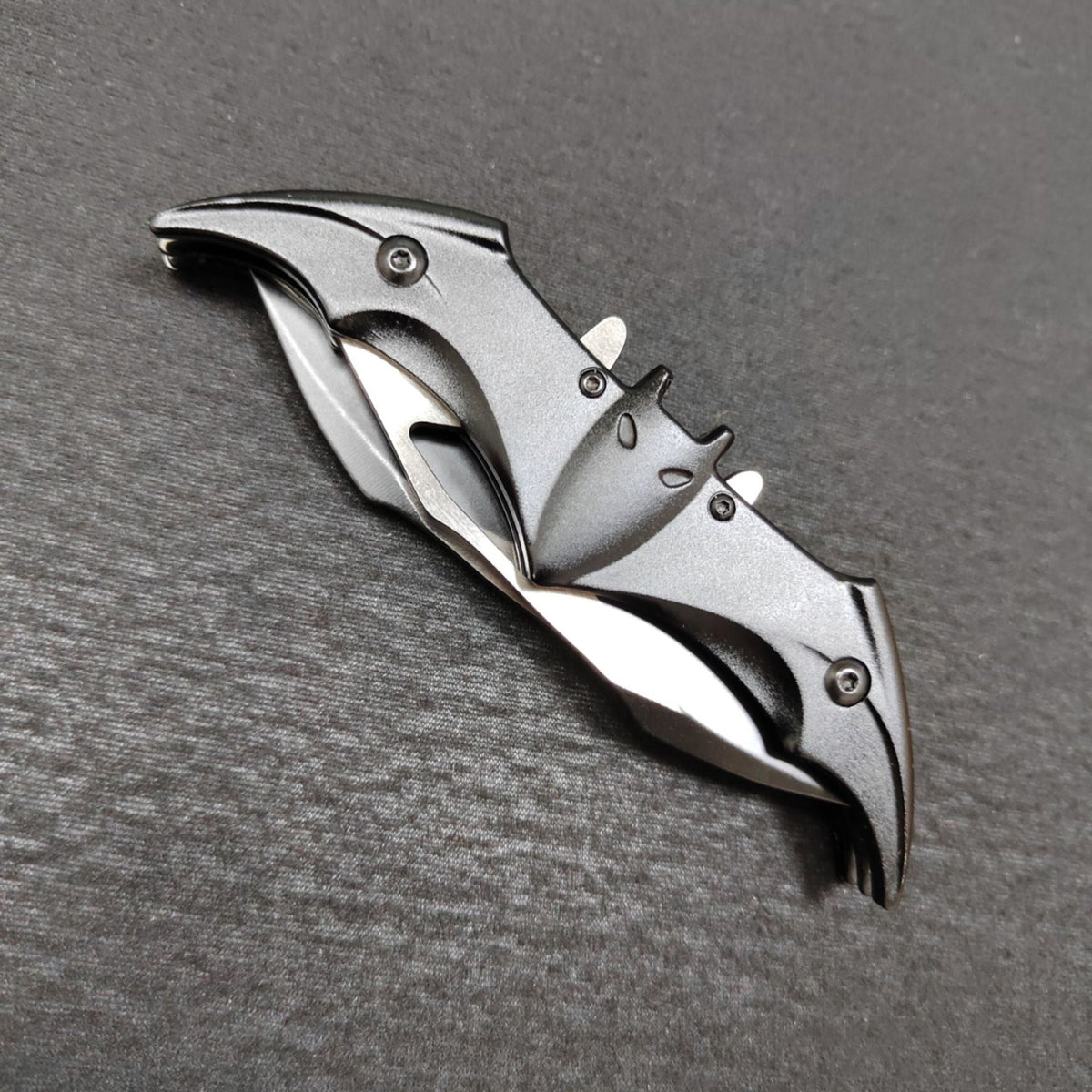 A closed black bat-shaped folding knife on a dark gray surface, its black bat handle showing ears, eyes and slotted edges with silver steel wing edges folded along the sides.
