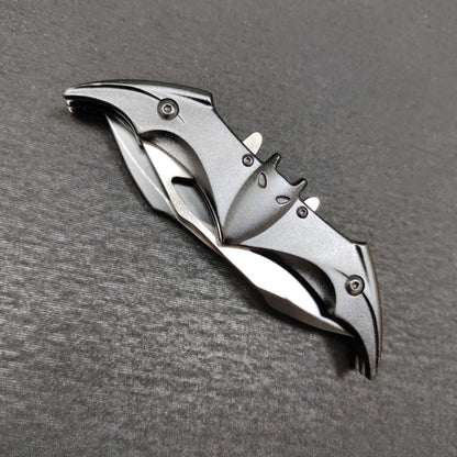 A closed black bat-shaped folding knife on a dark gray surface, its black bat handle showing ears, eyes and slotted edges with silver steel wing edges folded along the sides.