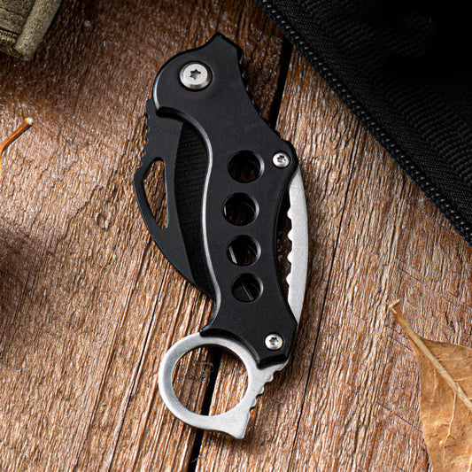 Closed Black Bird folding EDC claw knife with black handle resting on weathered wood next to a leaf.
