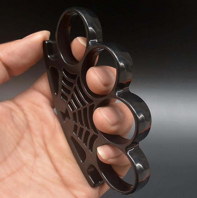 A black four-finger knuckle duster held in a hand, with four large oval finger holes and a spider-web pattern with a spider cast into the palm rest.