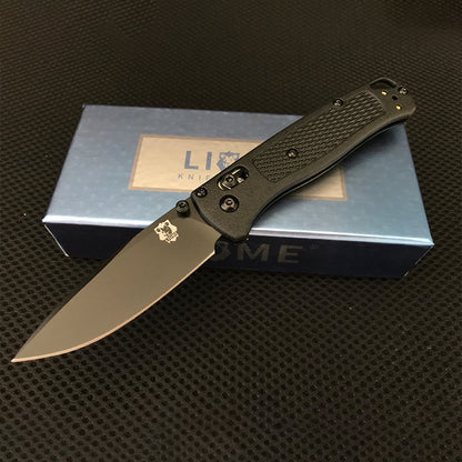 Black handled folding knife with black-coated drop-point blade and gold rear hardware open on a Liome box