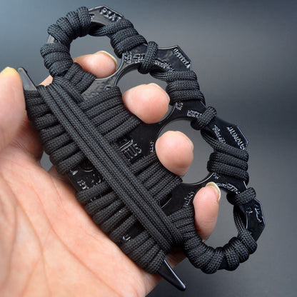 A hand holds a black zinc alloy knuckle duster wrapped in black paracord, its four finger holes fitted over the fingers and Latin words such as FIAT and MUNDUS embossed on the dark knuckle faces, against a dark gray background.