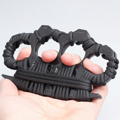 A hand holds a black four-finger knuckle duster wrapped in thick black paracord, the zinc alloy body showing embossed lettering along the outer edges, against a plain white background.
