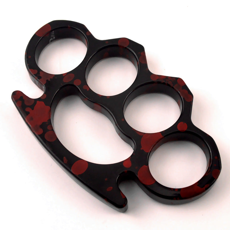 A glossy black knuckle duster with red splatter paint lying diagonally on a plain white studio background.