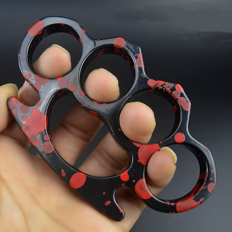 A glossy black knuckle duster with red splatter paint held in a hand, fingers through the four holes for size comparison, on a dark blurred background.