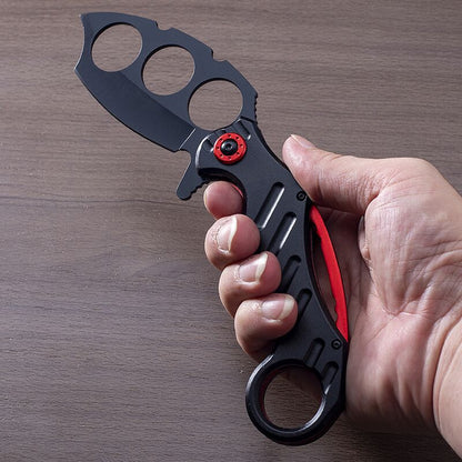 Black folding knuckle knife with red accents held in hand on wood surface