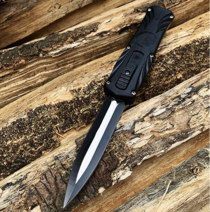 Black automatic OTF pocket knife with dagger blade extended on wooden logs