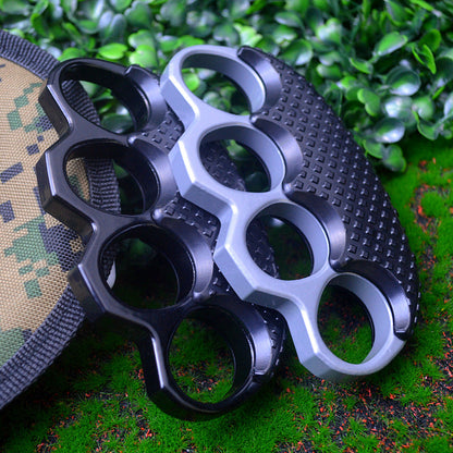 Black and silver gray knuckle dusters stacked together on a waffle-textured black case beside a camouflage pouch on green grass