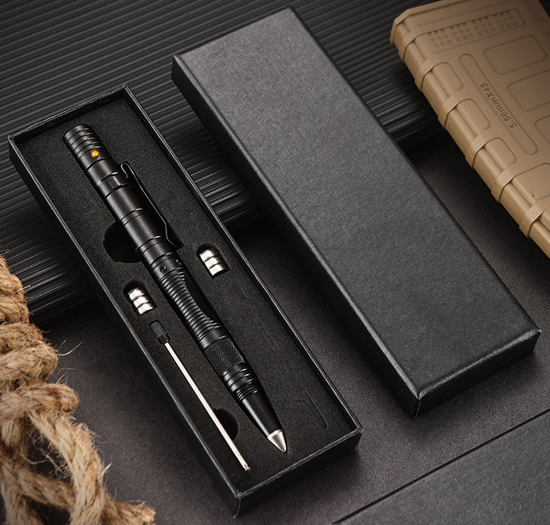 An open black gift box holding a black tactical pen with spare button batteries and a refill in the foam insert, set on a dark ribbed surface beside a coiled rope and a tan MOLLE pouch.