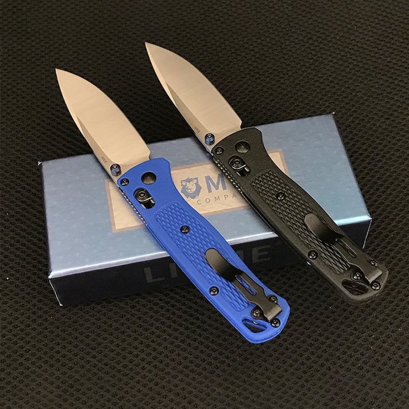 Blue and black handled folding knives with silver blades propped open against a Liome box, pocket clips facing outward