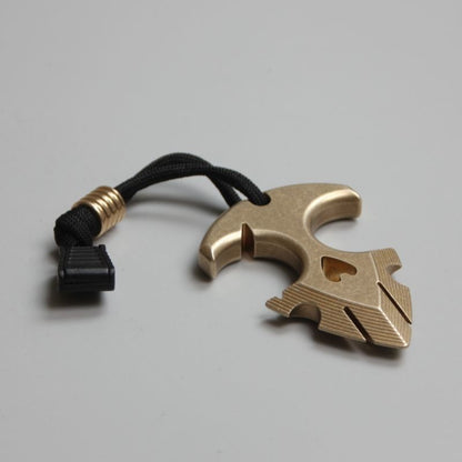 Brass anchor pendant with its black lanyard and cord lock resting on a dark gray surface, lit from the side.
