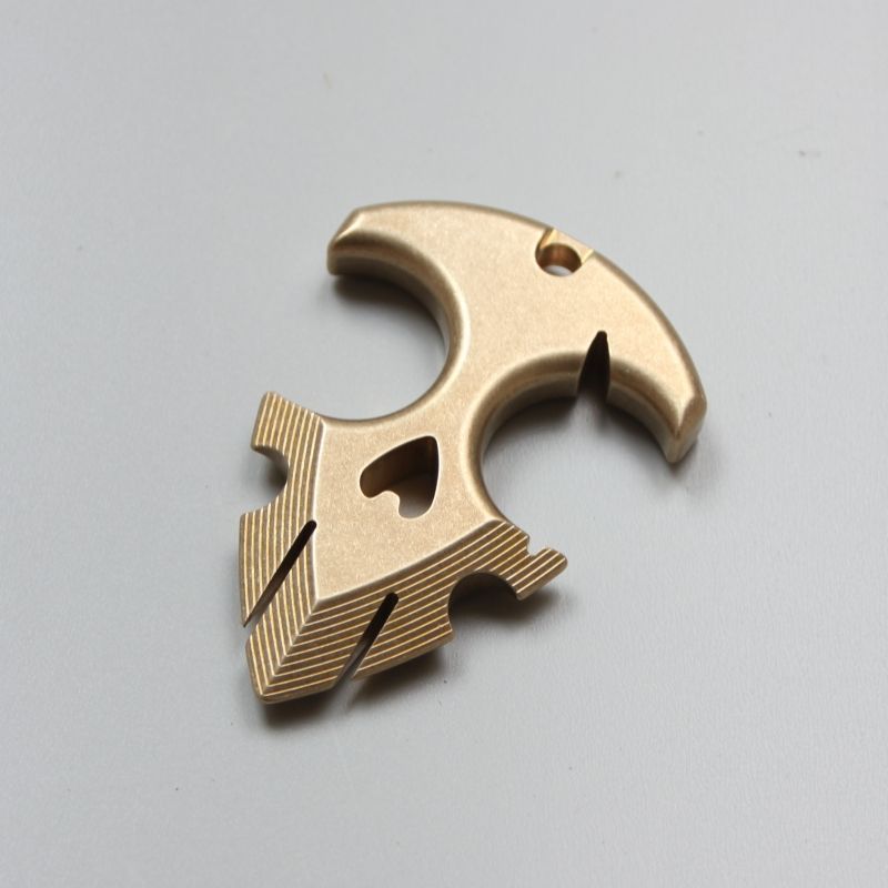 Brass anchor pendant lying on a light gray surface, showing the heart-shaped cutout and the ridged flukes.