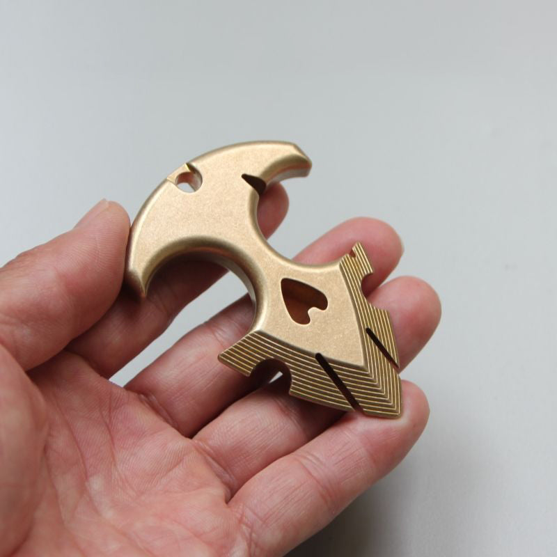 Brass anchor pendant held between the fingers, showing the heart-shaped cutout and stepped ridges on the flukes against a pale background.