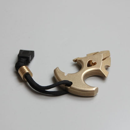 Brass anchor pendant seen from a low angle on a pale surface, with the black paracord looping around its shank.