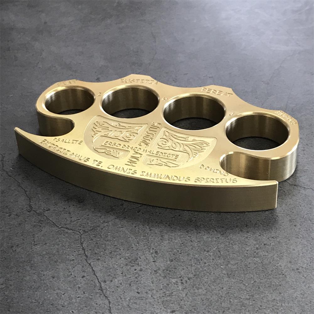 Low side angle of a brass knuckle duster on grey stone showing the long curved bottom edge with engraved lettering