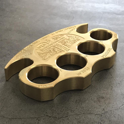 Engraved brass knuckle duster angled on dark grey stone with the claw tip pointing to the lower left