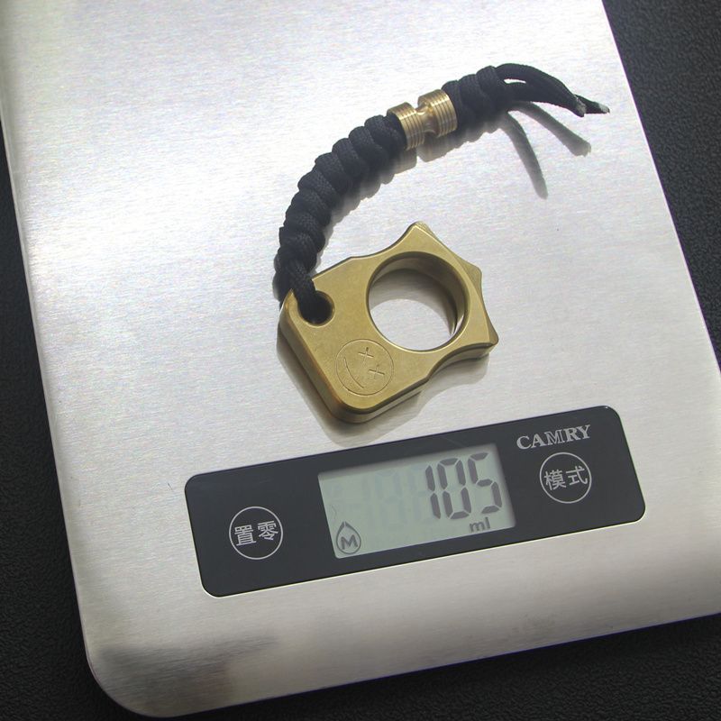 Brass knuckle duster on a digital scale reading 105, showing its weight with the lanyard attached.