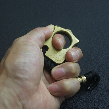 Hand gripping the brass knuckle duster with the index finger through the single hole and the lanyard hanging below.