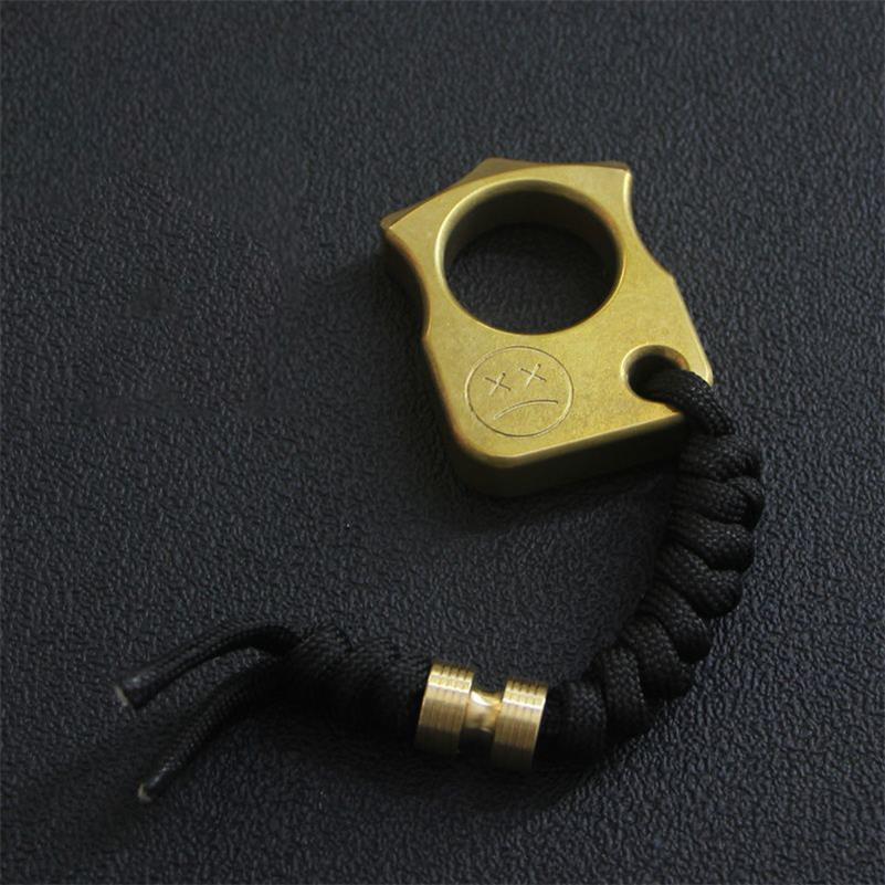 Single hole brass knuckle duster with a black paracord lanyard, showing the crossed-eye face engraving and a warm aged brass finish.