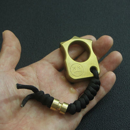 Palm-sized brass knuckle duster resting in an open hand, with its black paracord lanyard trailing across the palm.