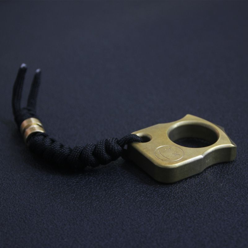 Side-angle view of the brass knuckle duster showing its thickness and edge profile, with the black lanyard stretched out to the left.