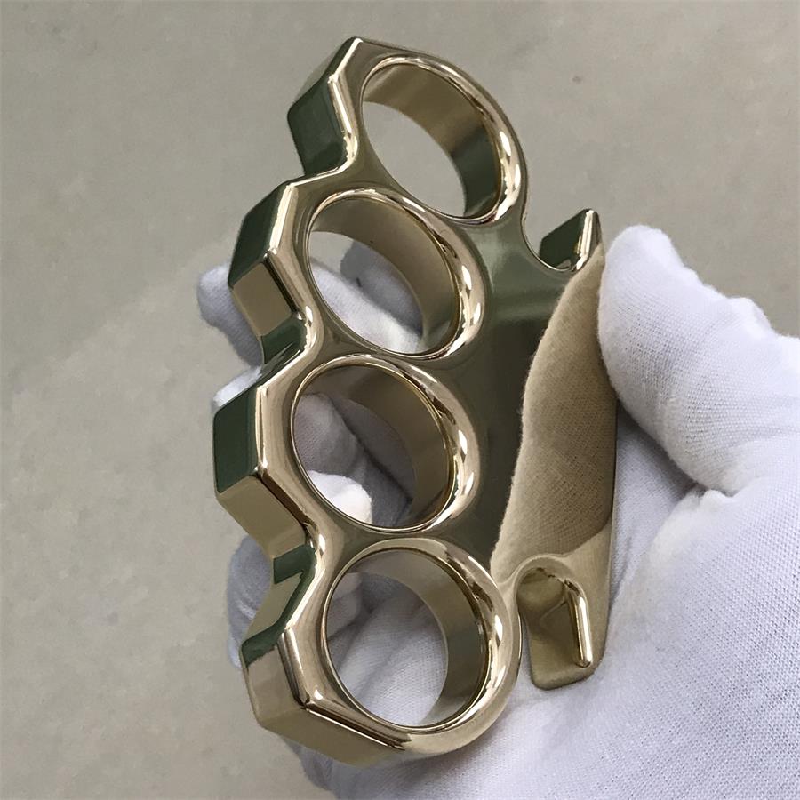 Brass knuckle duster held at a side angle showing the thick frame wall and rounded finger hole rims