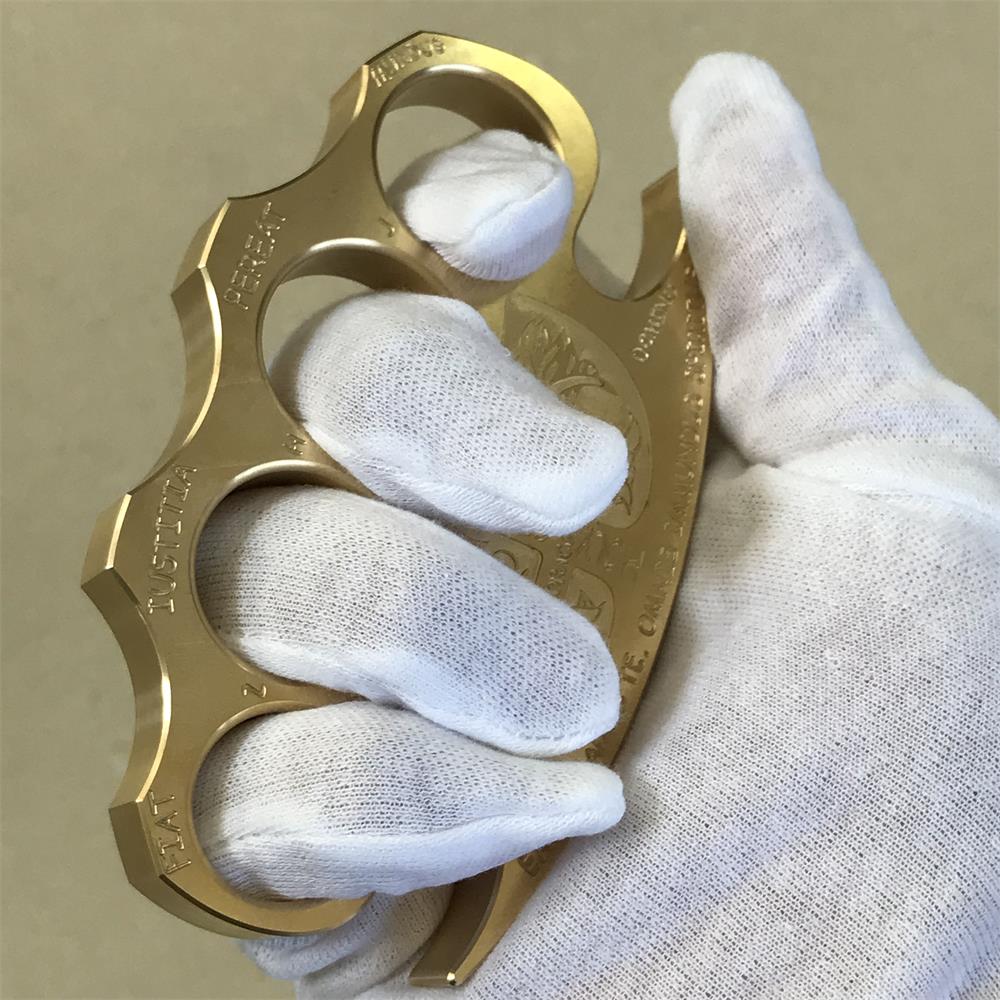 Vertical front view of an engraved brass knuckle duster held in a gloved hand, crest centered between the holes