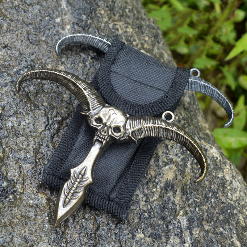 Bronze sheep skull pendant with a hanging ring on a black nylon sheath and silver horns behind among rocks and greenery