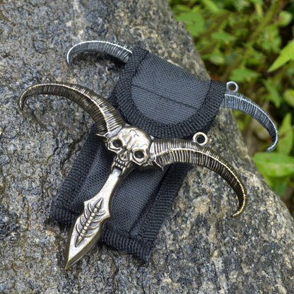 Bronze sheep skull pendant with a hanging ring on a black nylon sheath and silver horns behind among rocks and greenery