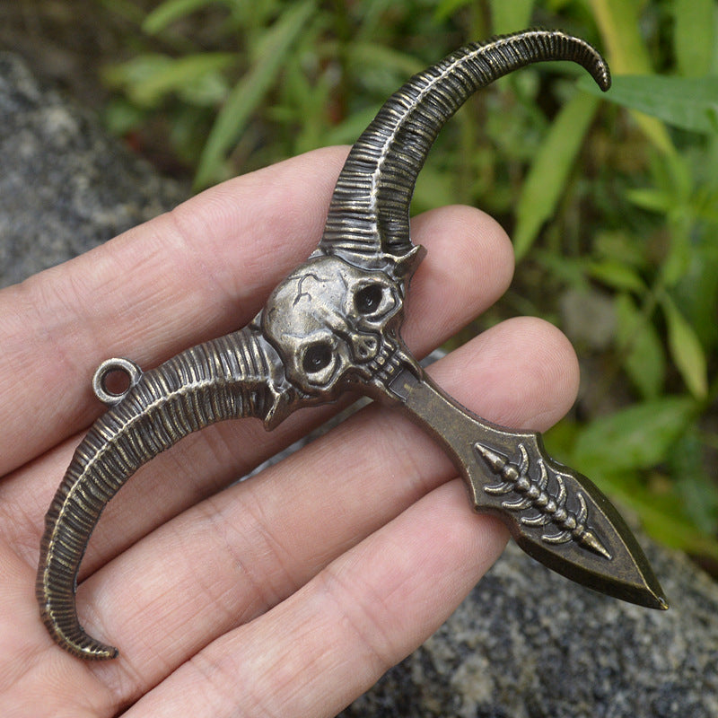 Bronze sheep skull pendant held in a hand for scale with ribbed horns and a hanging ring visible outdoors