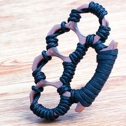Bronze woven knuckle duster angled on a wooden table with black umbrella rope around the finger openings and palm base